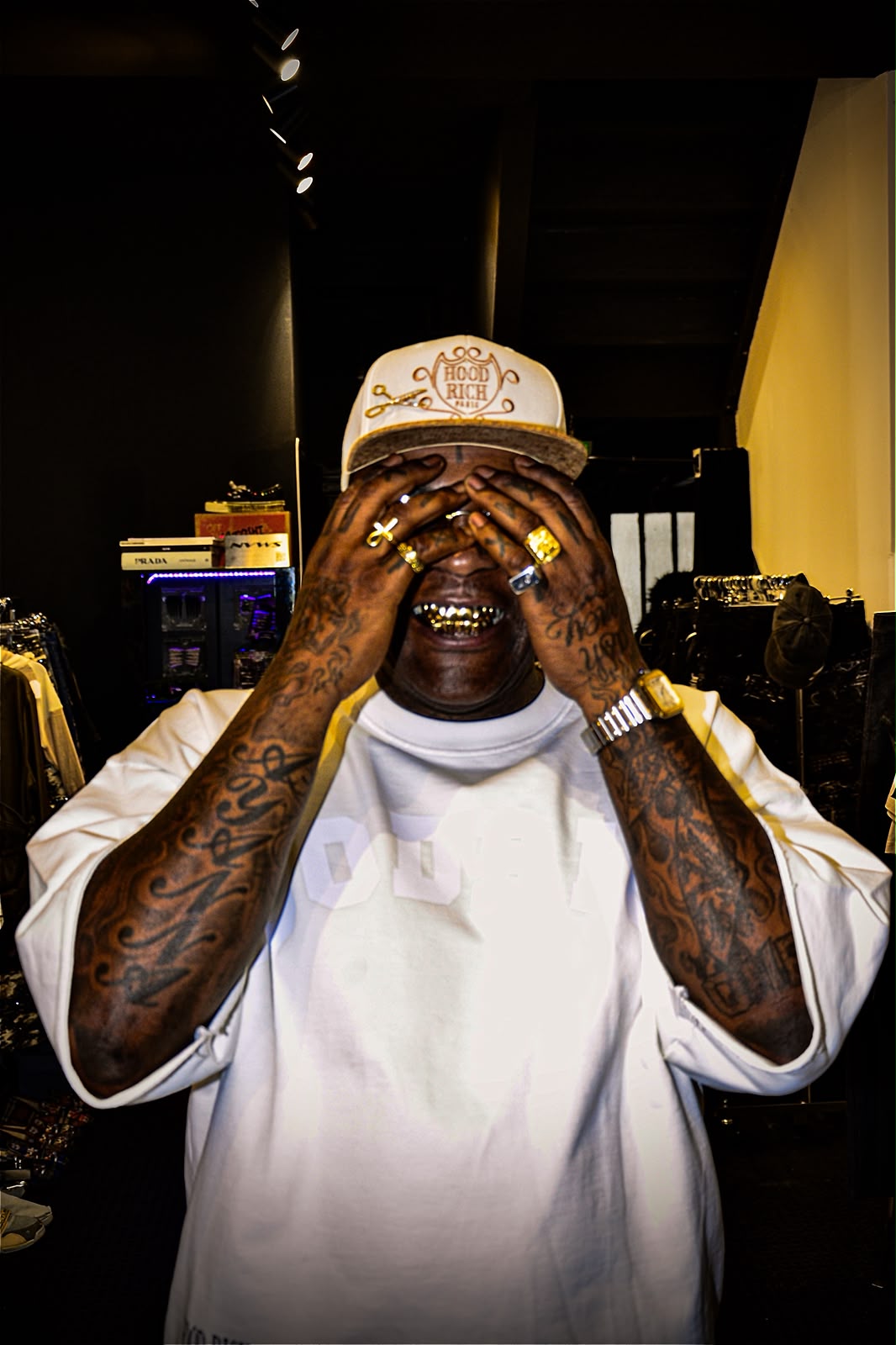 Homme en casquette blanche et t-shirt ample se couvrant les yeux de ses mains couvertes de tatouages et de bagues dorées, dans une boutique de vêtements