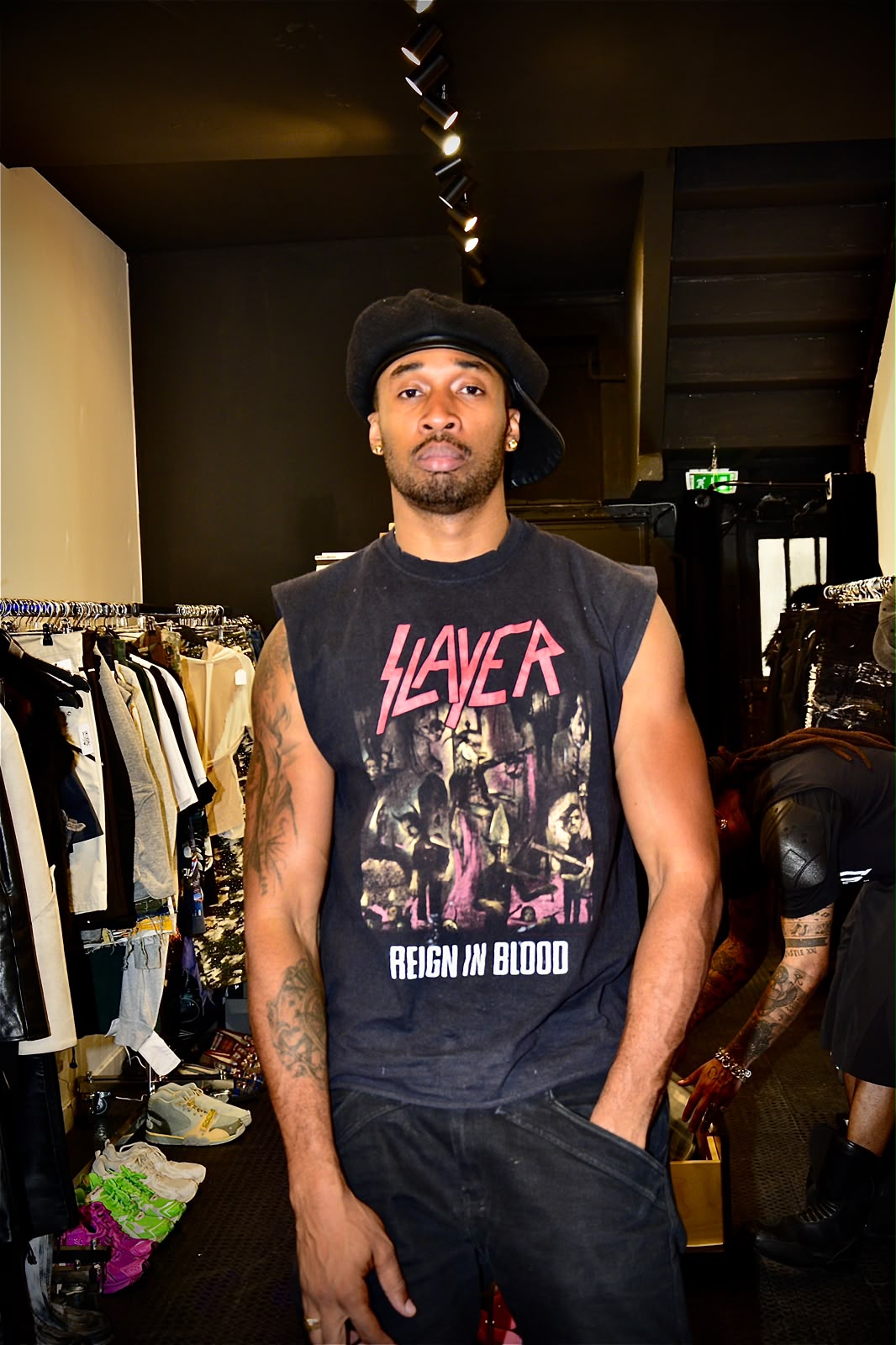 Homme en casquette noire et t-shirt de groupe sans manches posant entre les portants d'une boutique vintage