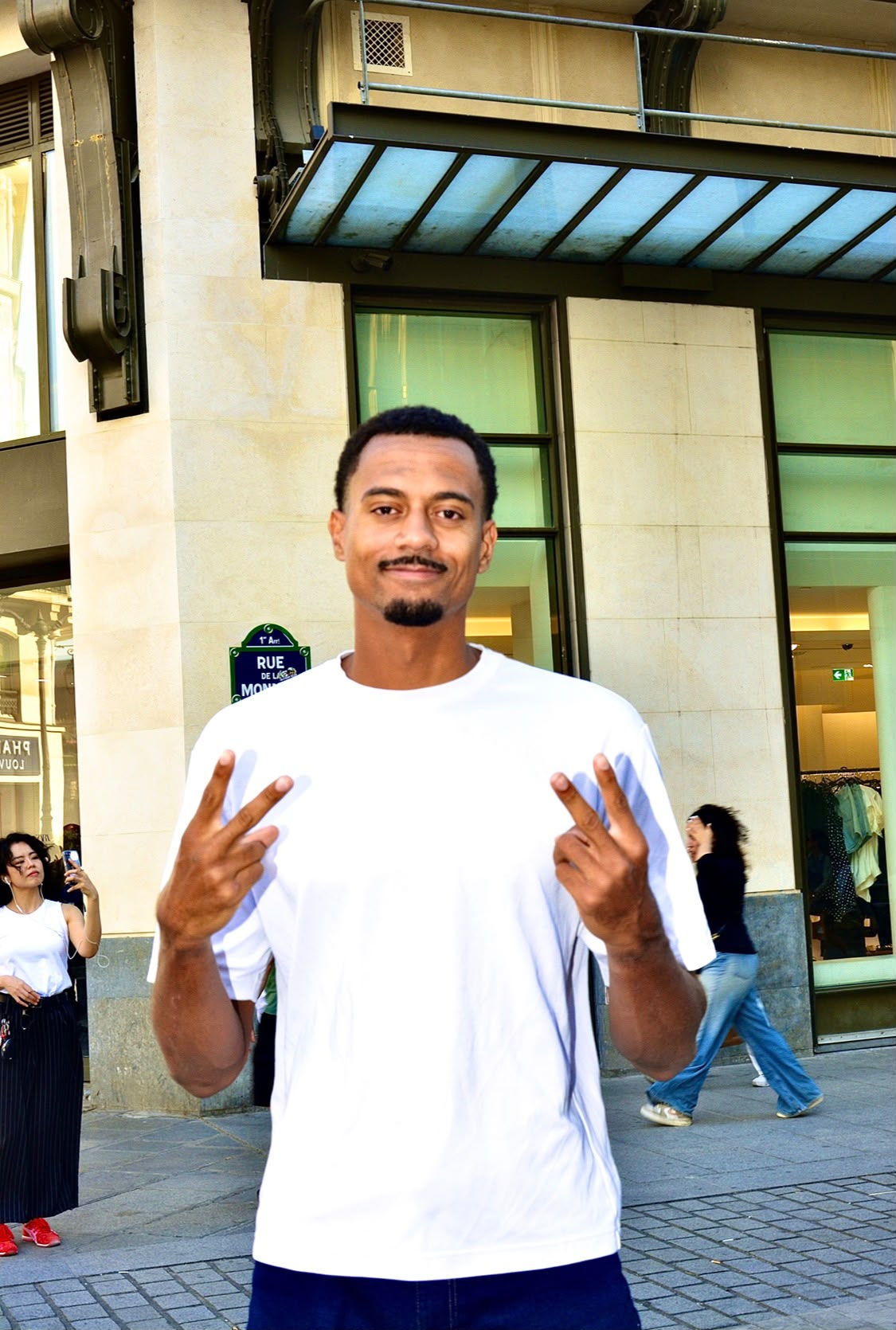 Homme en t-shirt blanc ample faisant le signe de la paix des deux mains rue de la Monnaie, à Paris