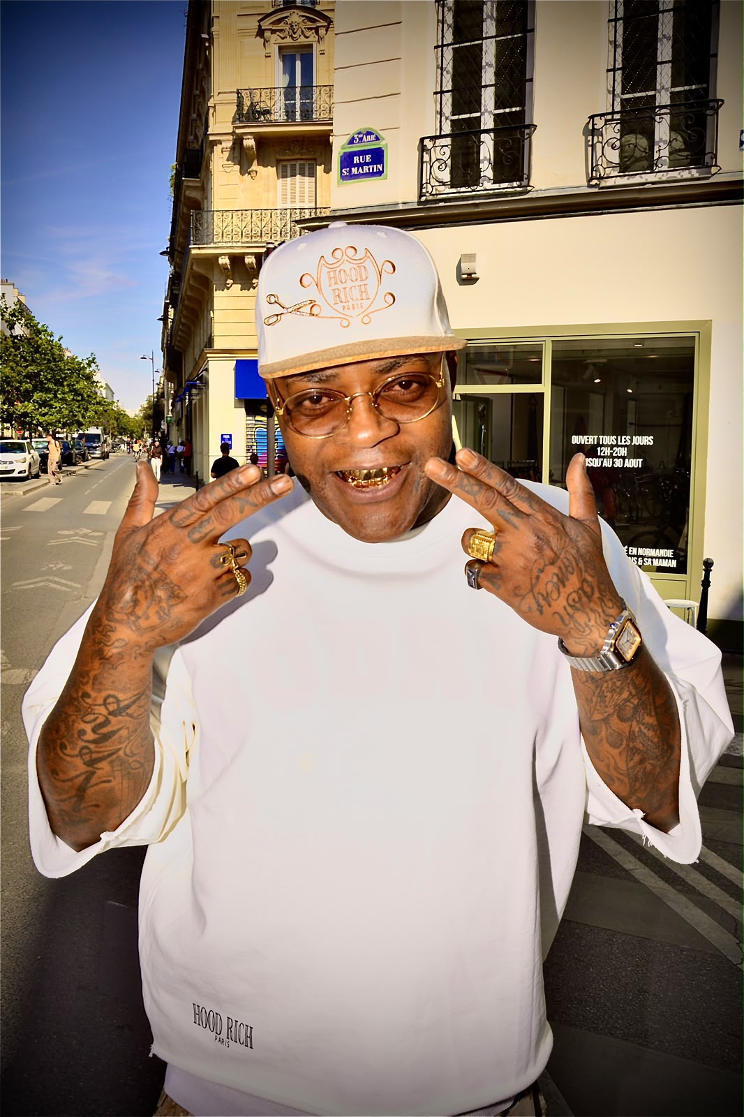 Gros plan d'un homme souriant en casquette blanche et lunettes dorées, bagues et tatouages aux mains, dans une rue de Paris