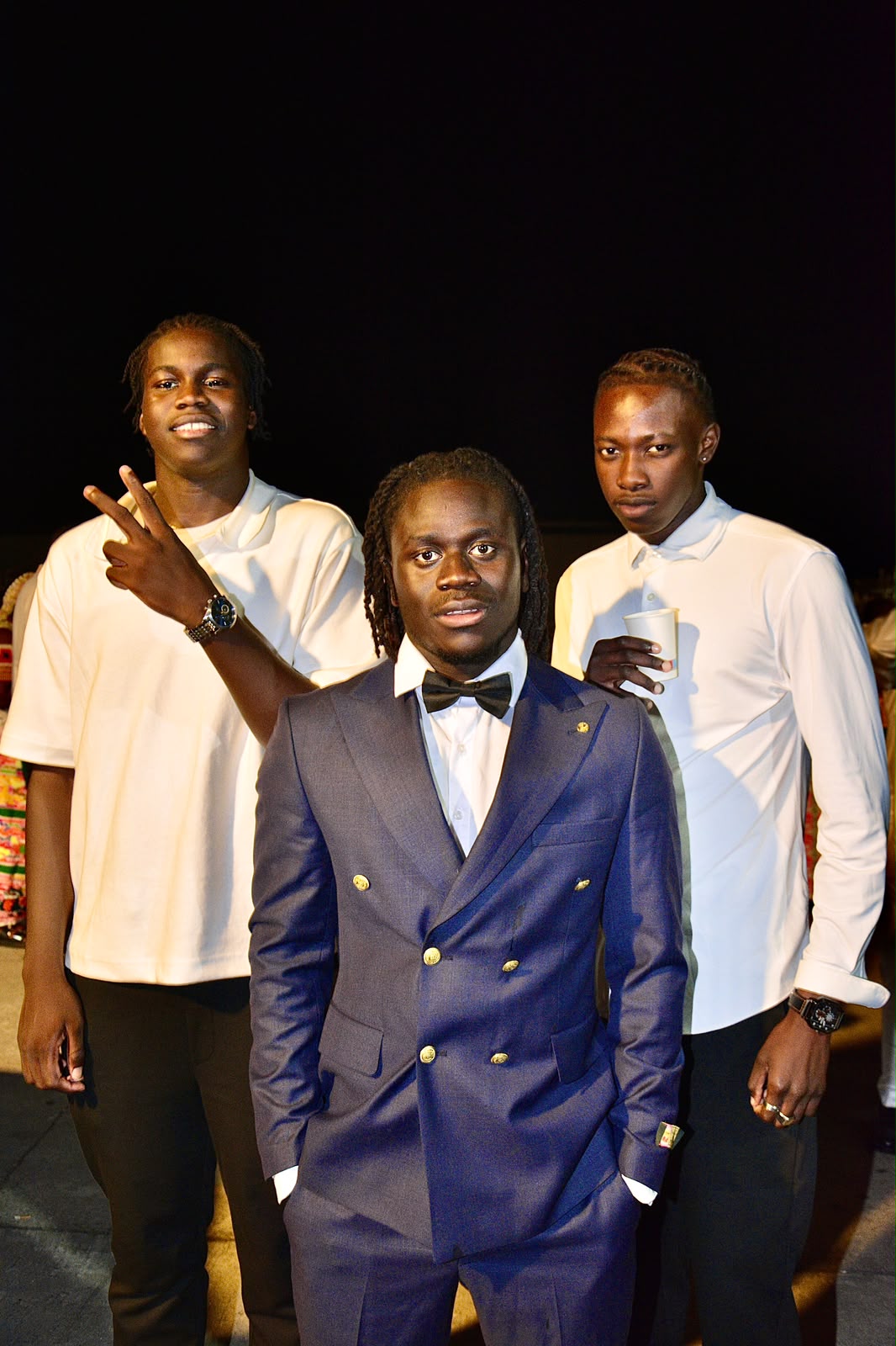 Trois jeunes posent la nuit ; celui du centre porte un costume bleu foncé avec nœud papillon, les deux autres une chemise blanche