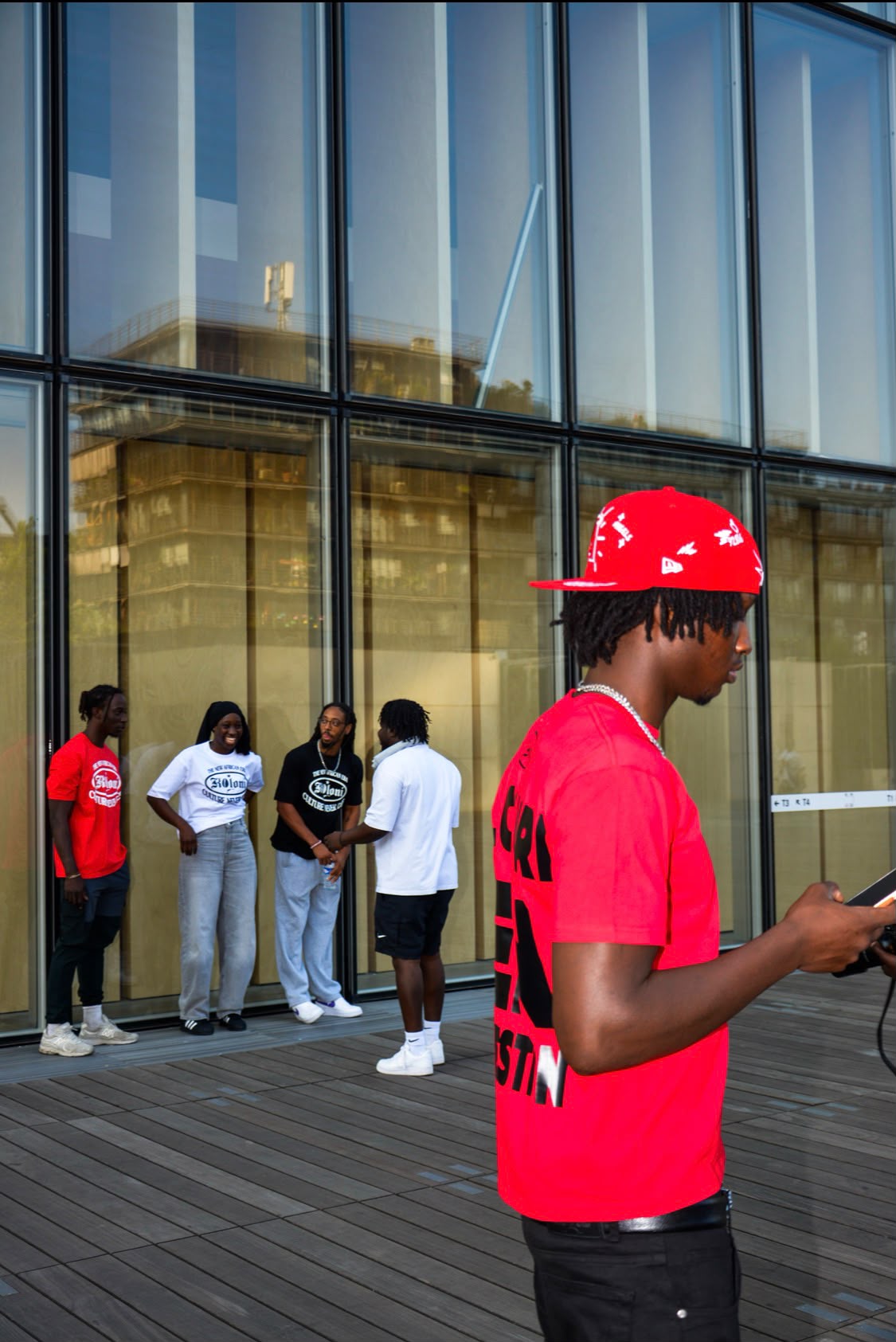 Jeune homme en t-shirt et casquette rouges de profil, avec un groupe reflété dans la façade vitrée en arrière-plan